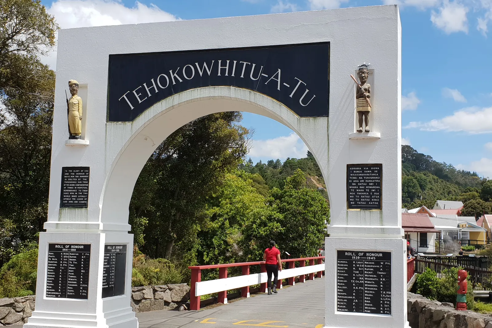 The white memorial gateway at the entrance to Whakarewarewa village, with a footbridge and a red-roofed house beyond