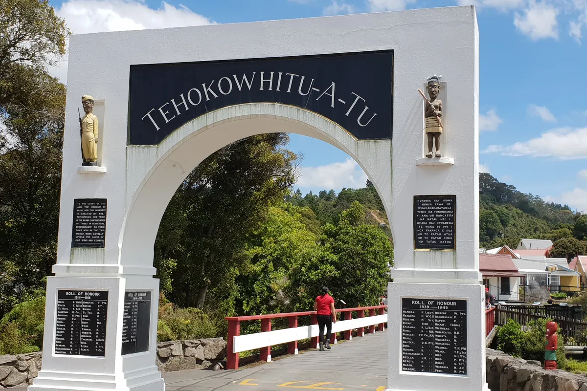 The white memorial gateway at the entrance to Whakarewarewa village, with a footbridge and a red-roofed house beyond