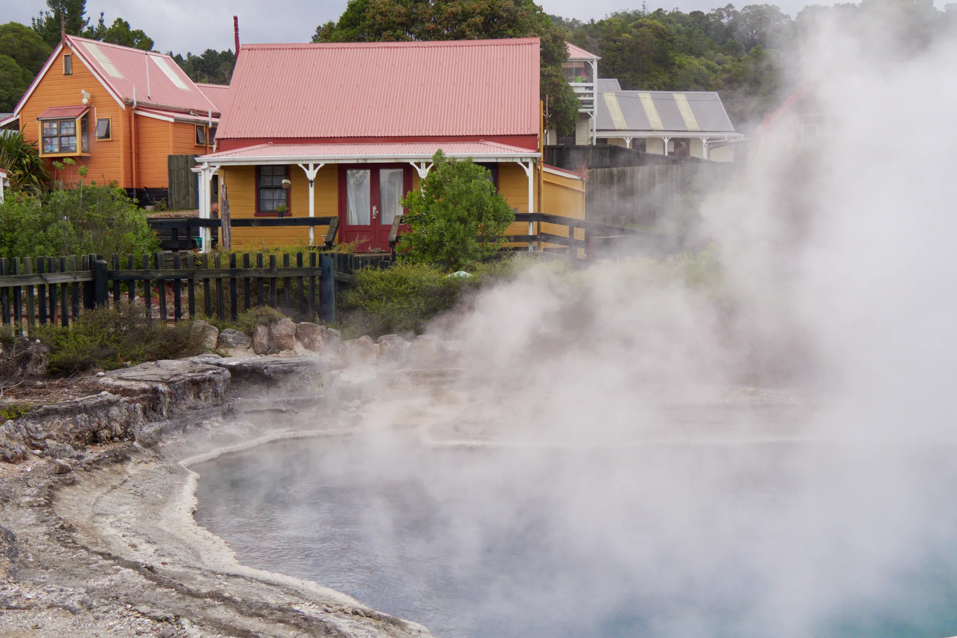 Yellow and orange wooden houses with red roofs stand behind a fence above a steaming blue-grey pool, with white steam rising across the right of the frame